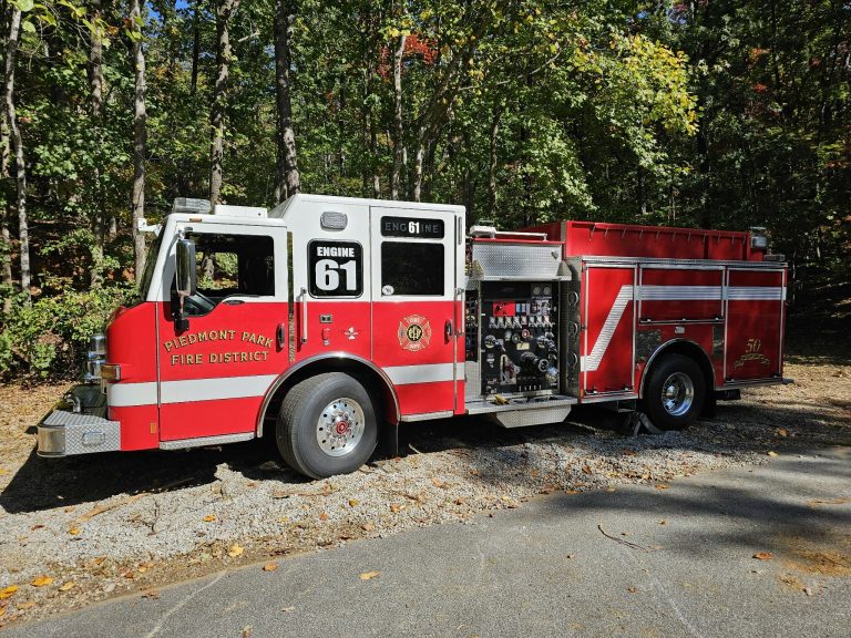 A red fire truck labeled "Engine 61" from the Piedmont Park Fire District is parked on a gravel path. It's surrounded by trees with autumn leaves.
