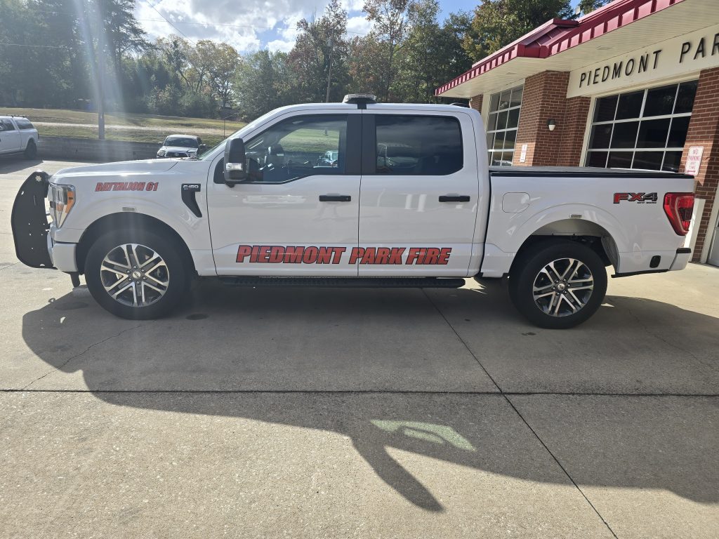A white pickup truck labeled "Piedmont Park Fire" is parked in front of a brick building. It has "Battalion 01" and "FX4" decals. The scene is sunny with trees and a lawn in the background.