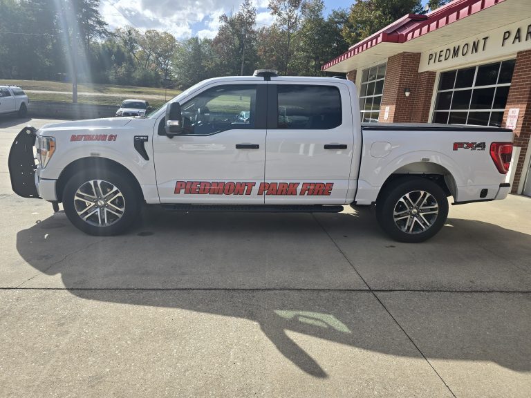 A white pickup truck labeled "Piedmont Park Fire" is parked in front of a brick building. It has "Battalion 01" and "FX4" decals. The scene is sunny with trees and a lawn in the background.