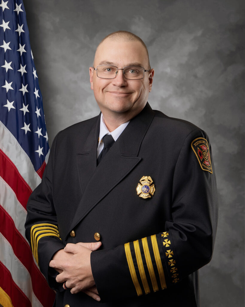 A man in a dark formal fire department uniform poses in front of an American flag, smiling at the camera. He wears glasses and has several gold stripes and badges on his jacket.