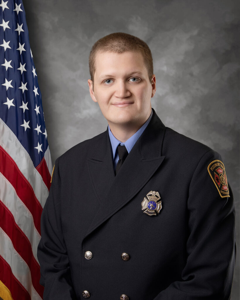 A person in a formal dark firefighter uniform stands in front of a U.S. flag and a mottled gray backdrop, looking at the camera with a slight smile. An embroidered fire department patch is visible on one sleeve.