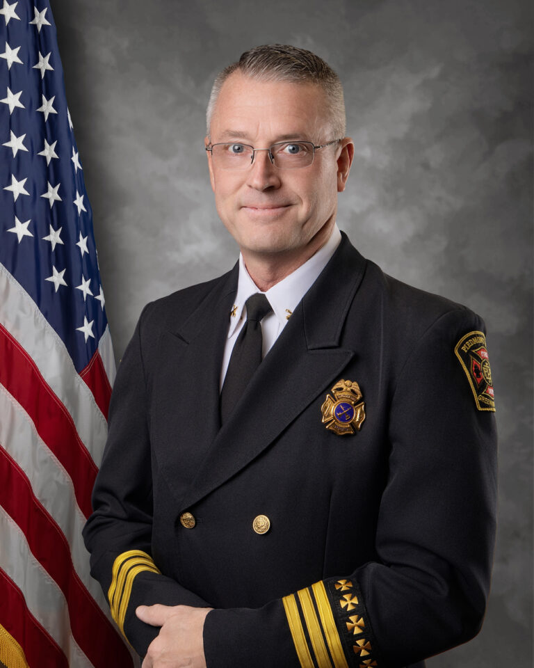 A man in a formal fire chief uniform stands in front of an American flag, smiling slightly. The uniform features gold buttons, a badge, and insignia on the sleeves and shoulders. The background is a neutral gray.