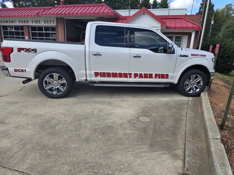 A white pickup truck with "Piedmont Park Fire" written on the side is parked in front of a building labeled "Piedmont Park Fire Dept." The truck has shiny chrome wheels and is parked on a concrete surface with grass nearby.