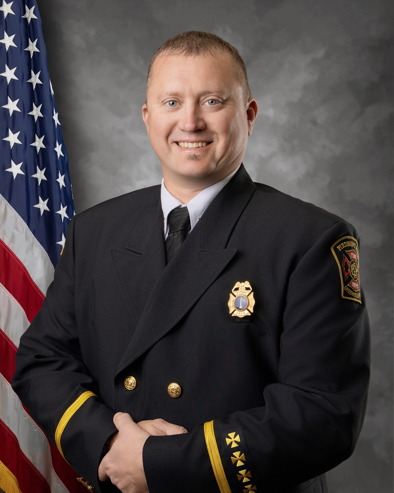 A smiling man in a dark formal uniform with badges stands in front of a gray backdrop and a U.S. flag. He has short blond hair and is posing with his hands clasped in front.