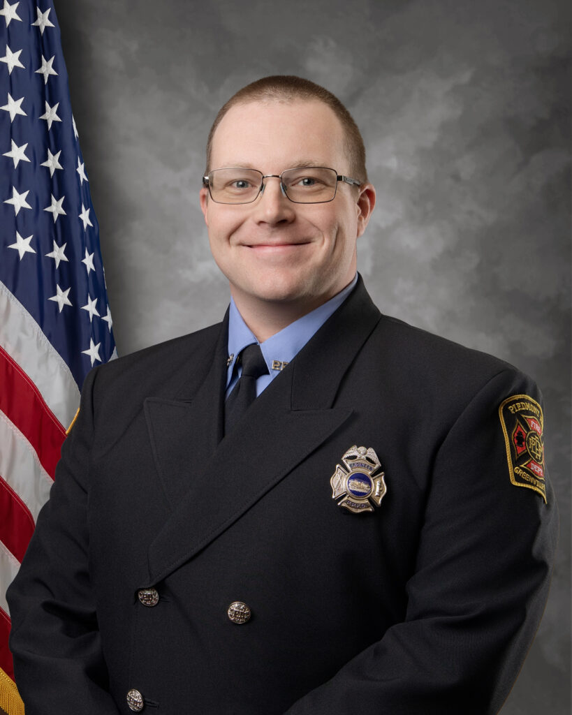 A smiling firefighter in formal uniform stands in front of an American flag and grey backdrop, with a badge and department patch visible on his jacket.