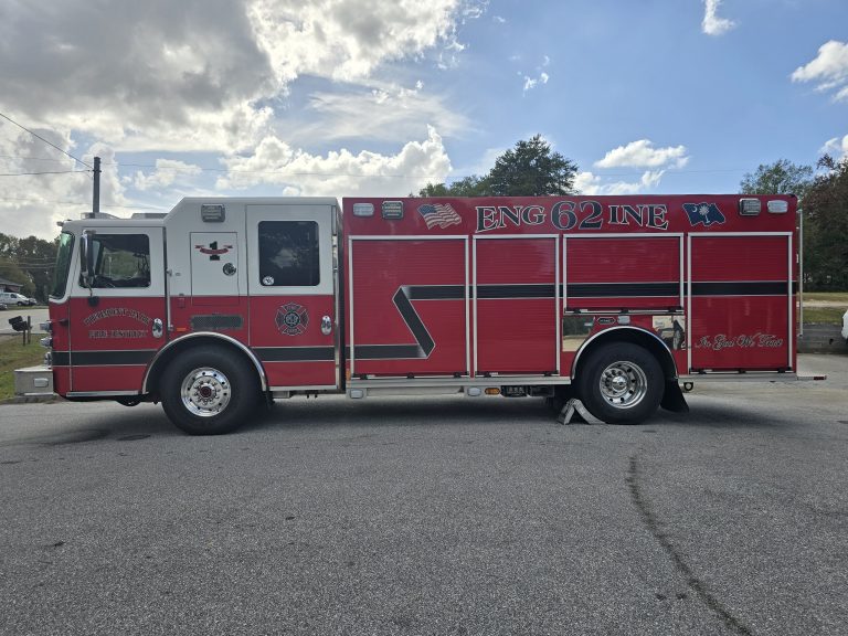 A red and white fire truck with "ENG 62" written on the side is parked on a paved area. The truck features various emblems and symbols, and the background includes a partly cloudy sky and trees.