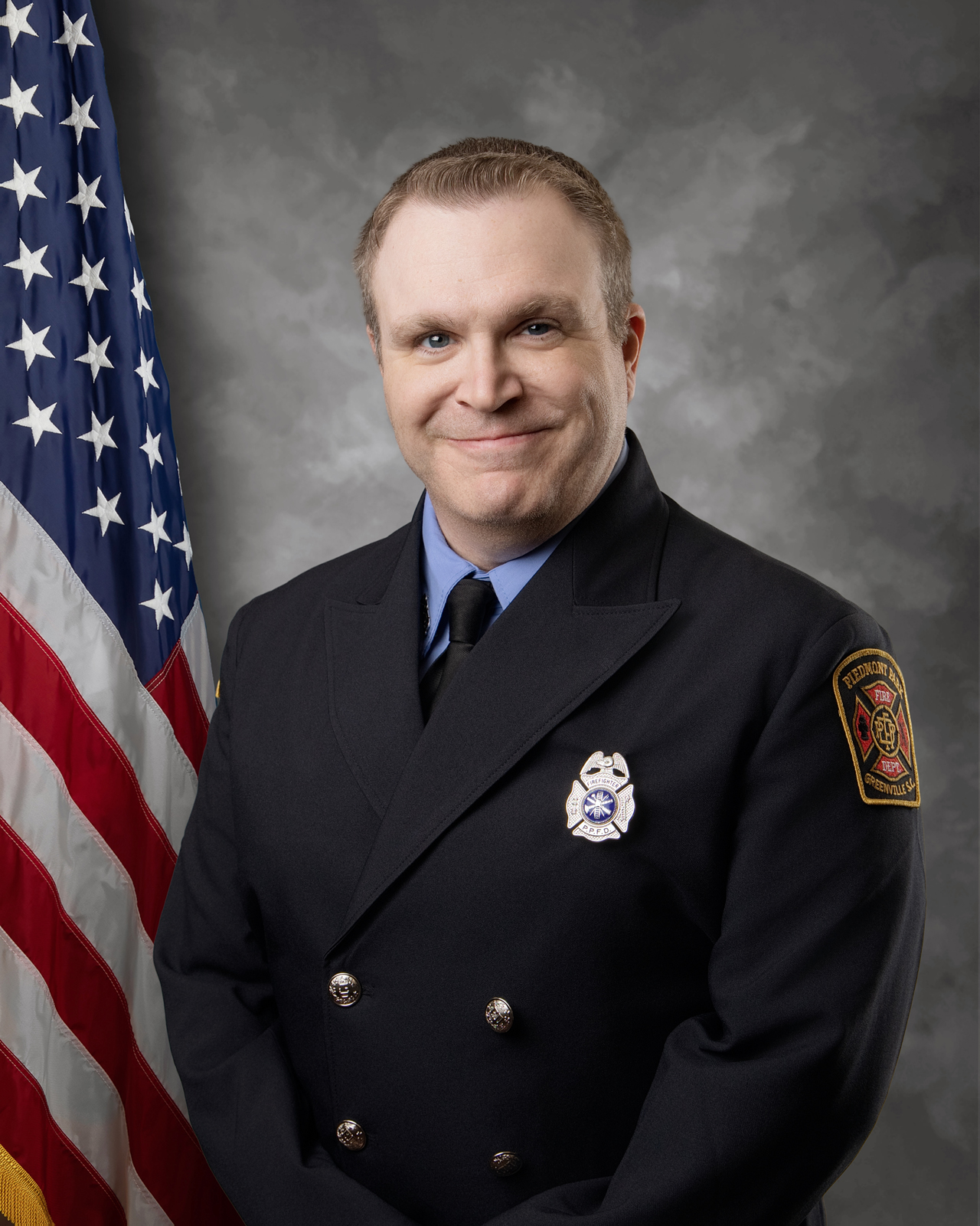 A firefighter in formal uniform smiles while standing in front of an American flag and a gray backdrop. He wears a badge and an insignia patch on his sleeve.