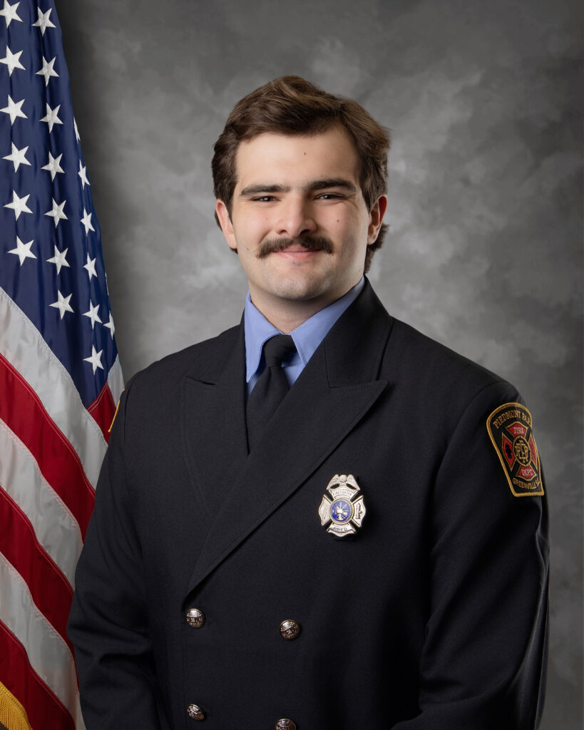 A firefighter in formal uniform stands in front of a US flag and gray backdrop, smiling slightly. He wears a double-breasted jacket with a badge and a patch on his shoulder.