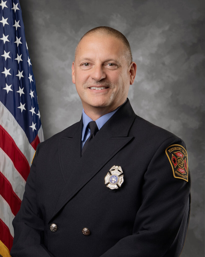 A firefighter in formal uniform stands smiling in front of an American flag and a gray background. He has short hair and wears a badge and a patch reading "Fire Department.