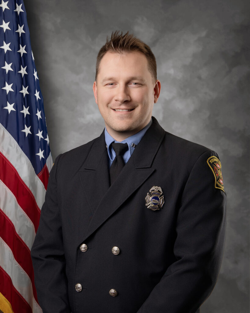 A male firefighter in dress uniform stands smiling in front of a gray backdrop and a U.S. flag. He has short hair and wears a badge on his coat.