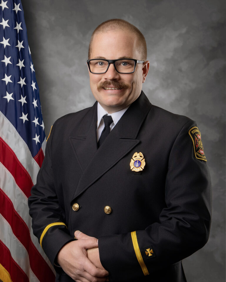 A man in a formal dark fire department uniform with a badge and insignia stands in front of an American flag, smiling with his hands clasped. He wears glasses and has a mustache.