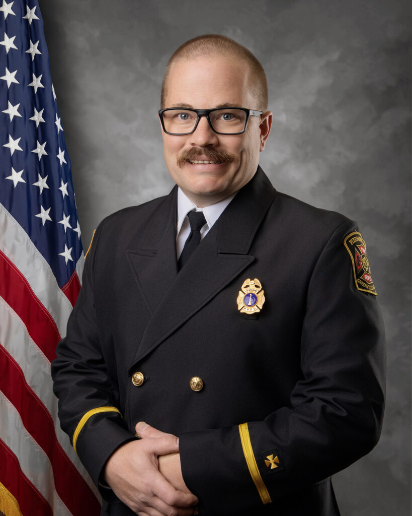 A man in a formal dark fire department uniform with a badge and insignia stands in front of an American flag, smiling with his hands clasped. He wears glasses and has a mustache.