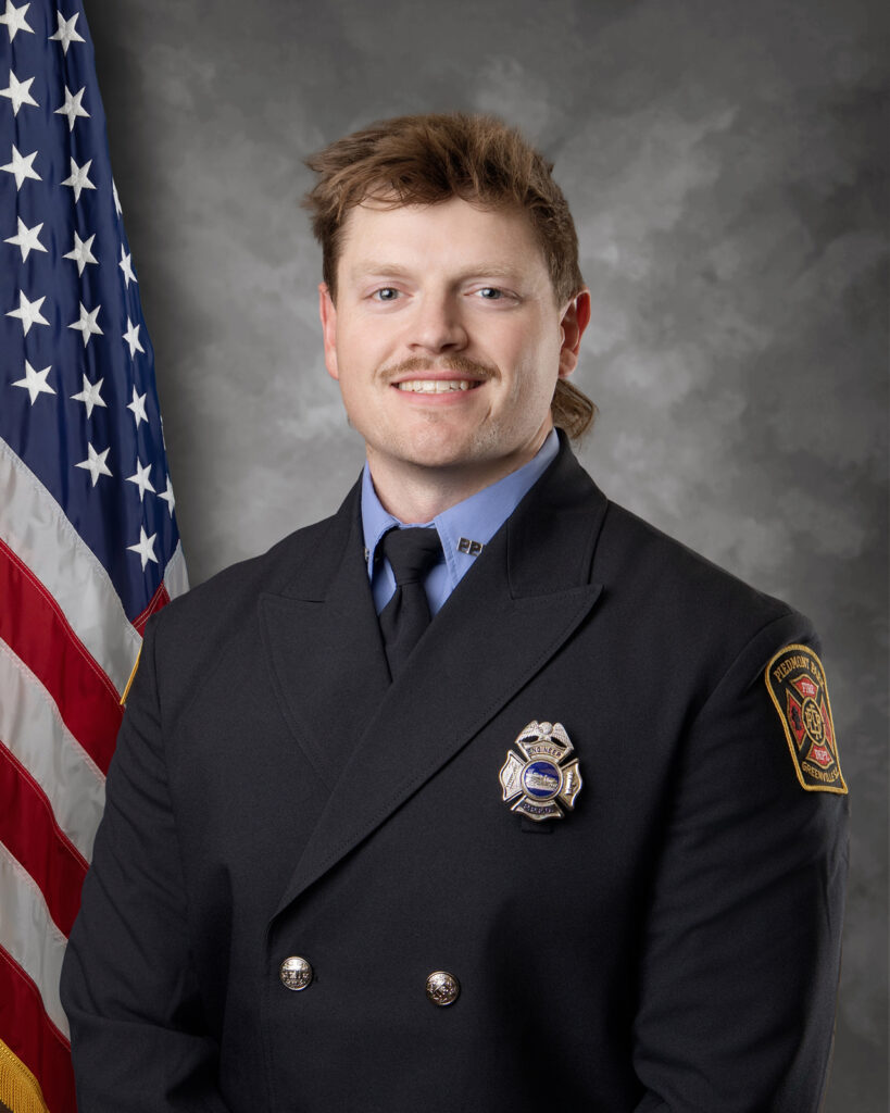 A firefighter in formal uniform stands in front of an American flag, smiling slightly. The uniform has a badge and a patch on the shoulder. The background is gray and mottled.