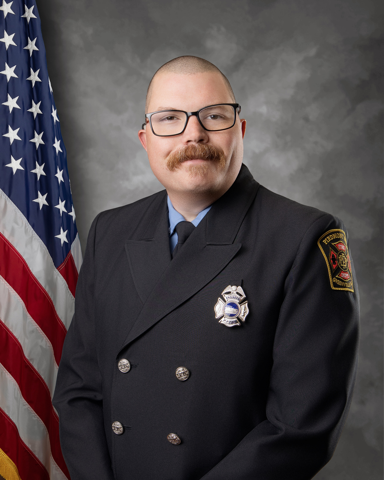 A firefighter in formal uniform stands in front of a gray backdrop and an American flag, wearing glasses and a badge, with a department patch on his shoulder and a mustache.