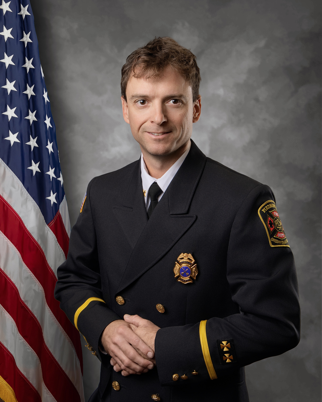 A man in a formal fire department uniform stands in front of an American flag, smiling, with hands clasped. He wears badges and insignia, indicating a high rank. The background is a neutral gray.