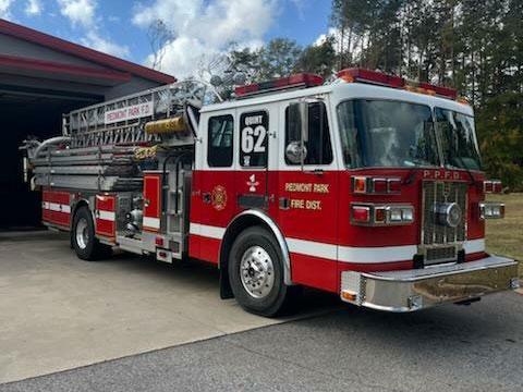 A red and white fire truck with a ladder parked outside a fire station. The truck is marked with "Unit 62" and "Piedmont Park Fire Dept" on its side. Trees and a cloudy sky are visible in the background.