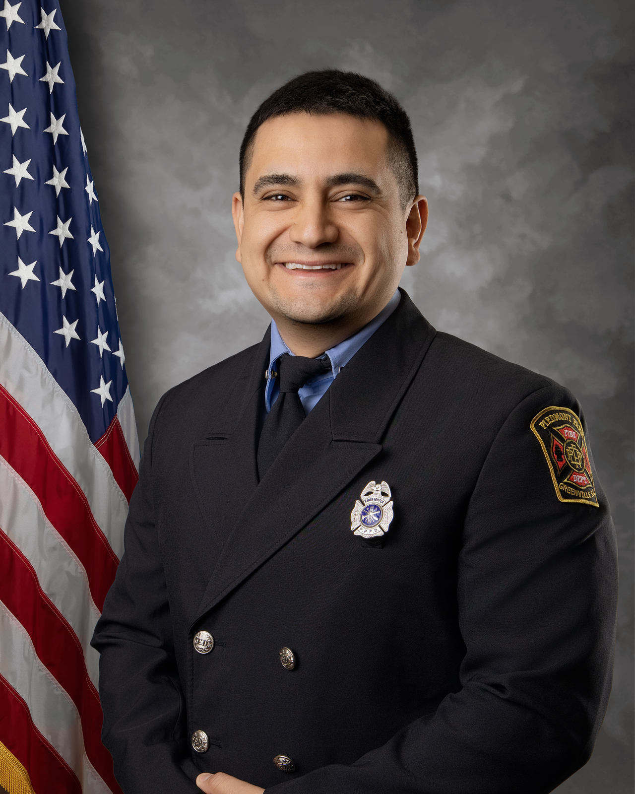 A smiling firefighter in uniform stands in front of an American flag, posing for a formal portrait with a gray backdrop. The badge and patch on the uniform are visible.