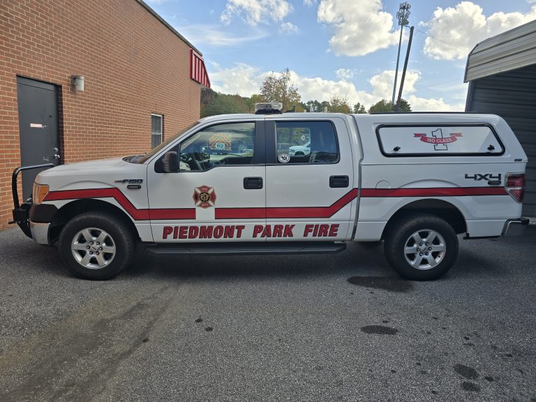 A white pickup truck with "Piedmont Park Fire" written on the side is parked on a paved surface. The truck has red and gold fire department logos and is parked next to a brick building. The sky is partly cloudy.