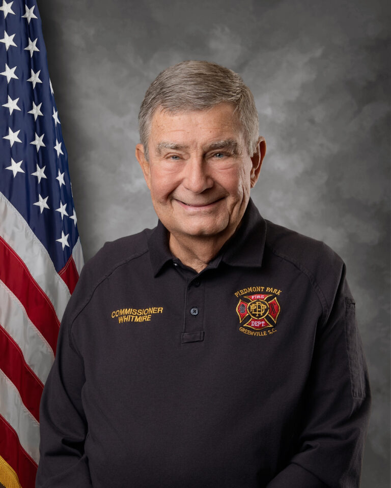 An older man with short gray hair, wearing a black Piedmont Park Fire Department shirt that reads "Commissioner Whitmire," stands smiling in front of an American flag and a gray backdrop.