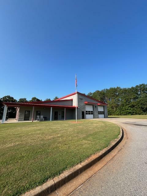 A single-story building with a red roof sits under a clear blue sky. It has a small porch, two large garage doors, and an American flagpole in front. A curved, paved driveway leads up to the building, which is surrounded by a well-maintained lawn and trees in the background.
