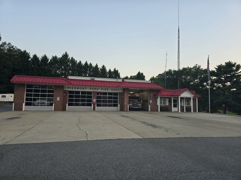 A fire station with a sign reading "Piedmont Park Fire Dept" on the building. The station has three large garage bays, two open and one closed. A fire truck is visible inside one of the open bays. A tall antenna and a flagpole with an American flag are on the right side.
