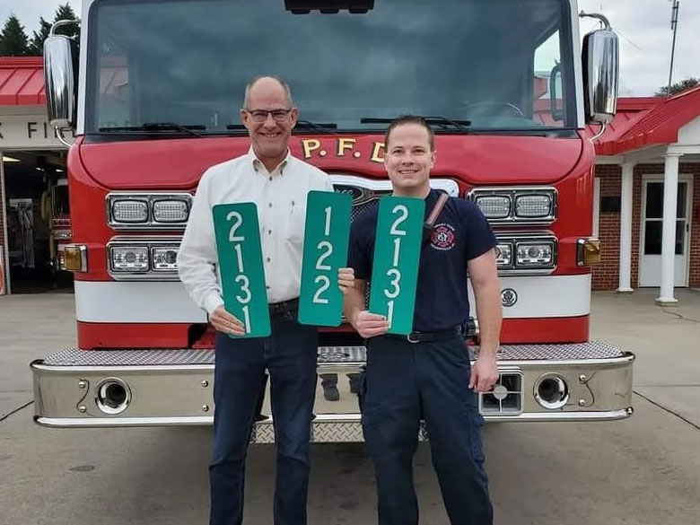 Two men stand in front of a red fire truck holding green address signs with numbers "2131" and "122." The man on the left wears a white shirt, and the one on the right is in a fire department uniform.