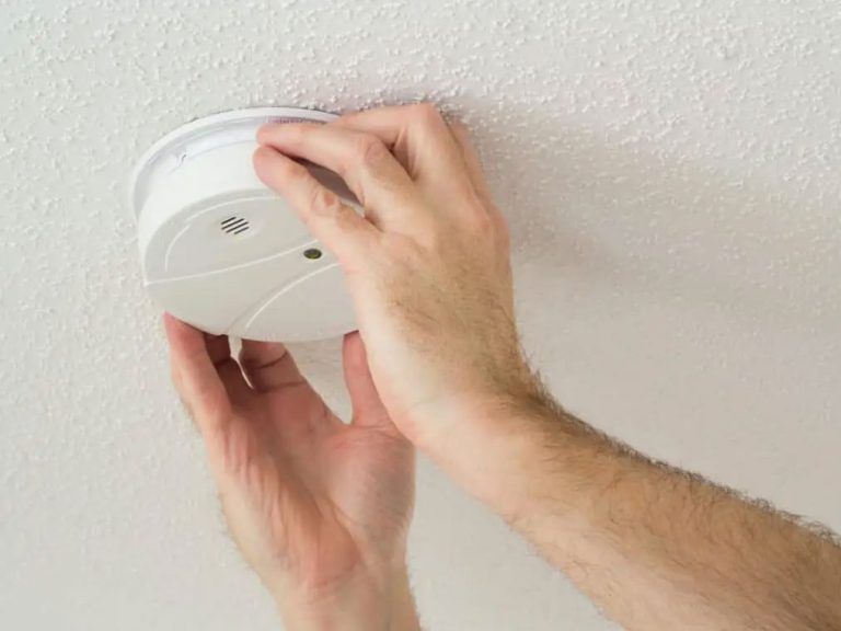 A person is installing or checking a white smoke detector on a textured ceiling. The person's hands are visible, adjusting the device, which is round and features a small circular opening and a green indicator light.