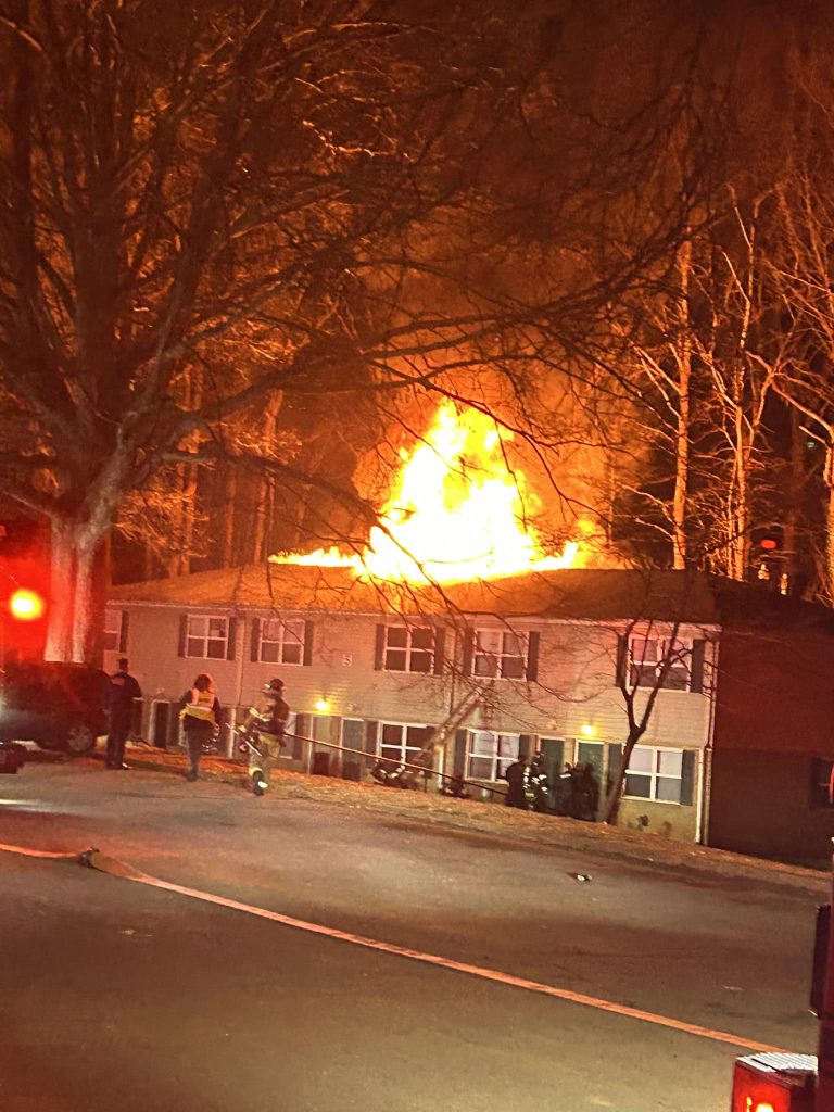 A large fire engulfs the roof of a multi-story building at night. Flames illuminate the scene, and emergency responders are visible nearby. A firetruck is parked on the street, casting red lights over the area. Trees surround the building.