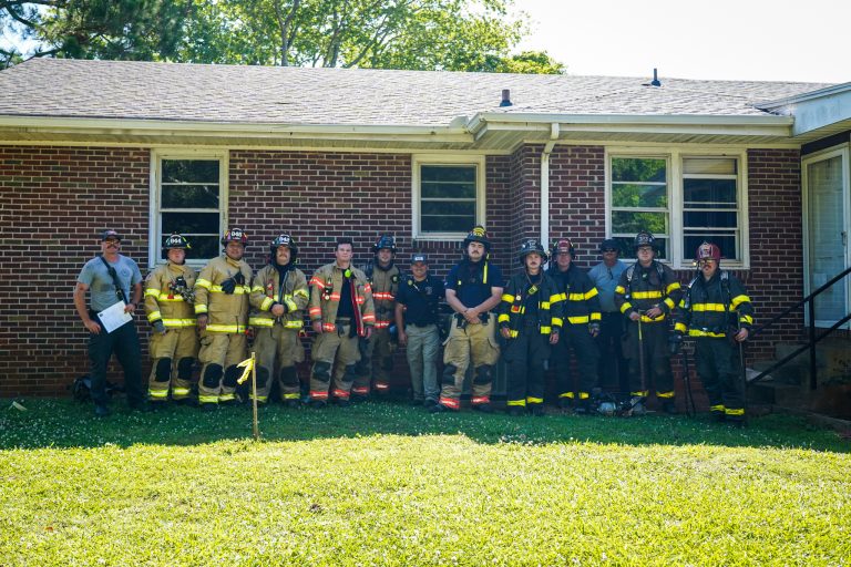 A group of firefighters in uniform stand in a row outside a brick house on a sunny day, posing for a group photo on the grass in front of the building.