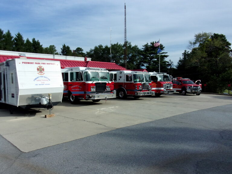 Four fire trucks and a fire department trailer are parked outside a fire station with a red roof. Trees and an American flag are visible in the background under a clear sky.