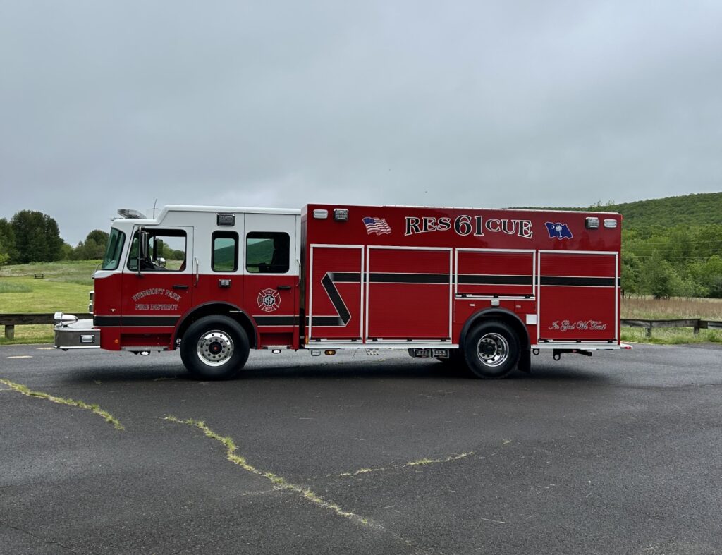 A large red rescue fire truck labeled "RESCUE 61" is parked on an empty paved lot with grassy fields and trees in the background under a cloudy sky.