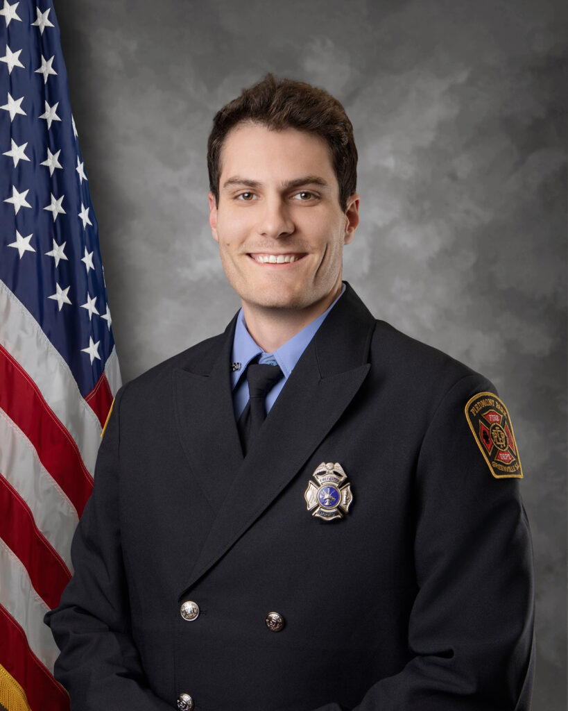 A firefighter in uniform stands smiling in front of a U.S. flag and a gray backdrop. He wears a dark jacket with a badge and an embroidered shoulder patch.