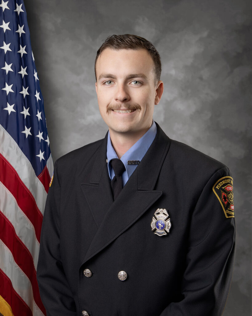 A firefighter in uniform with a badge poses for a formal portrait in front of a U.S. flag and a gray backdrop, smiling slightly at the camera.