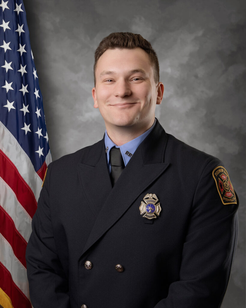 A smiling firefighter in uniform stands in front of a U.S. flag and a gray background. He wears a dark double-breasted jacket with a badge, a blue shirt, and a black tie.