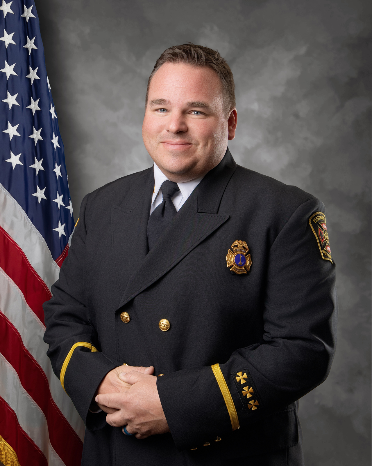 A man in a formal dark uniform with gold buttons and badges stands in front of an American flag, smiling and clasping his hands. He has short hair and a grey background behind him.