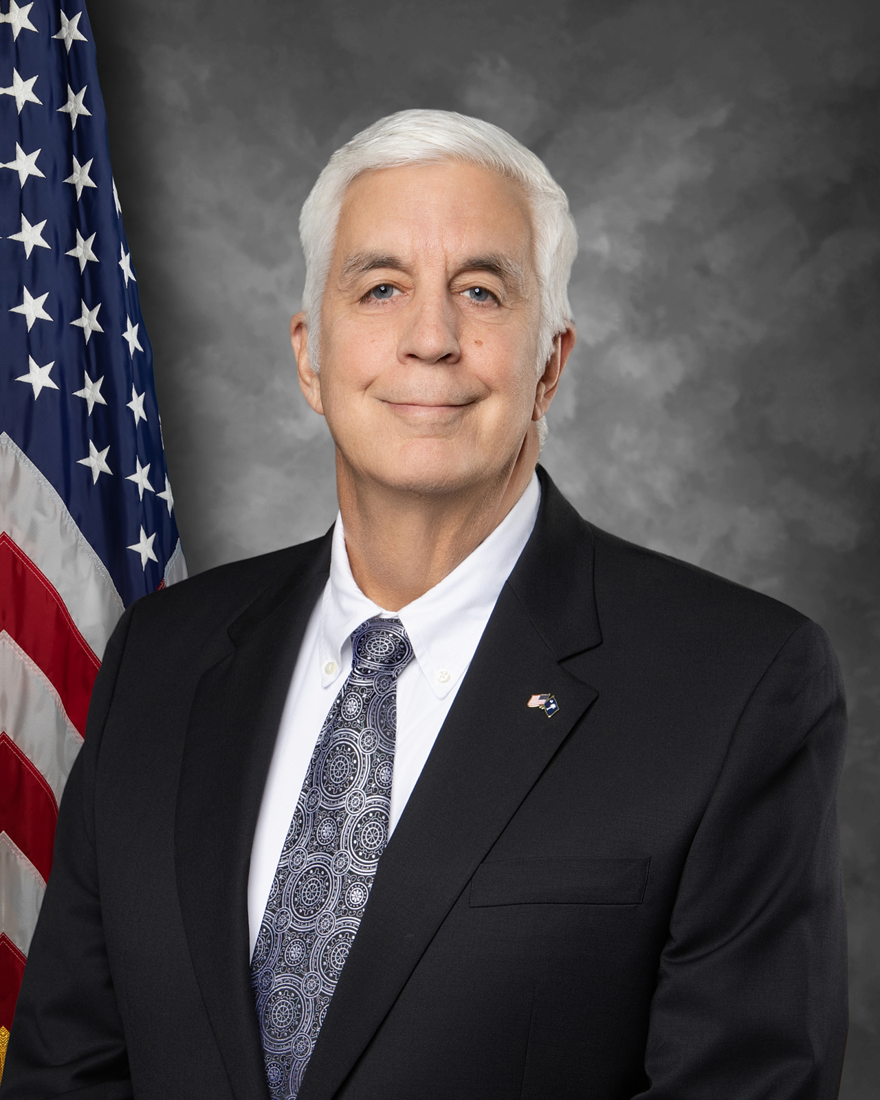 A man with short white hair in a dark suit, patterned tie, and white shirt stands in front of the American flag and a gray mottled background, smiling slightly at the camera.