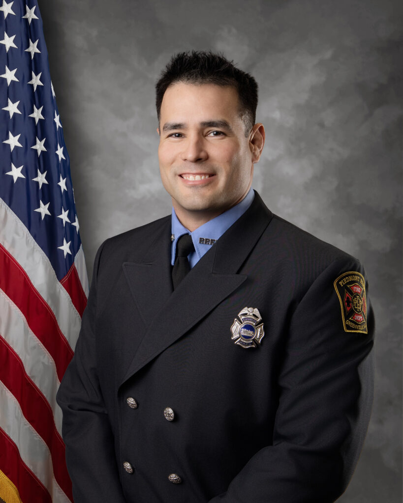 A firefighter in formal uniform stands in front of a gray backdrop and an American flag, smiling at the camera. The uniform features a badge, department patch, and double-breasted buttons.