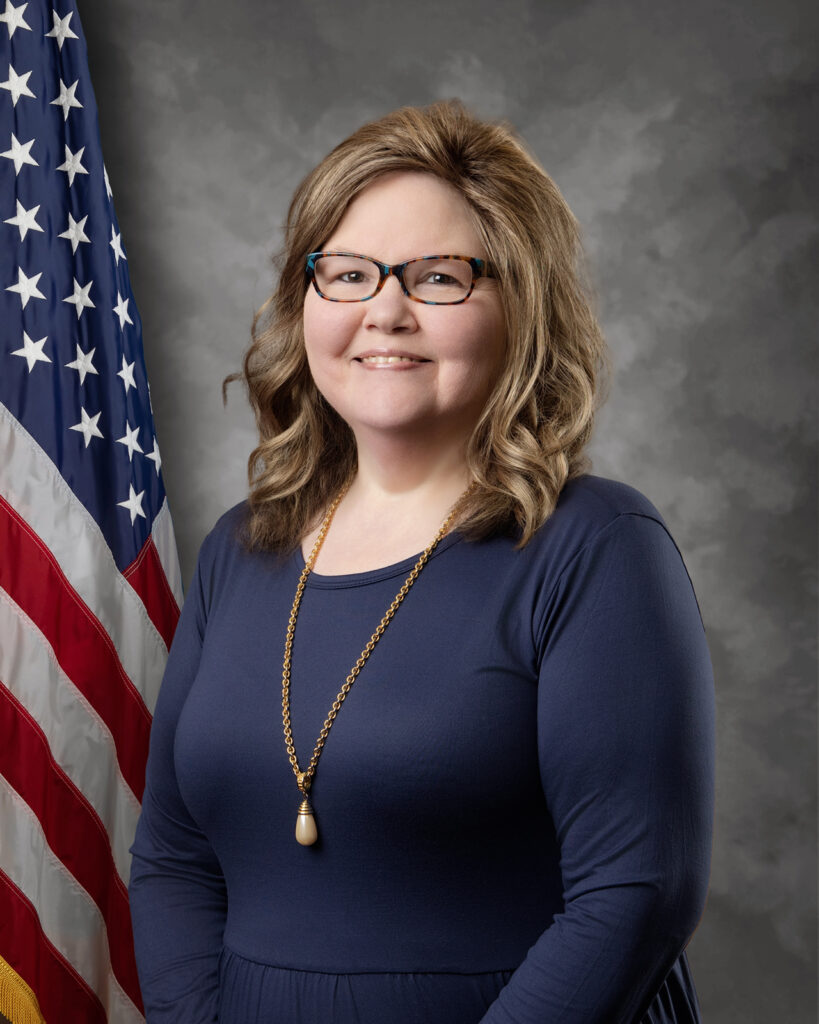 A woman with wavy light brown hair, glasses, and a navy blue dress stands in front of an American flag and a gray background, smiling at the camera. She wears a long gold necklace.