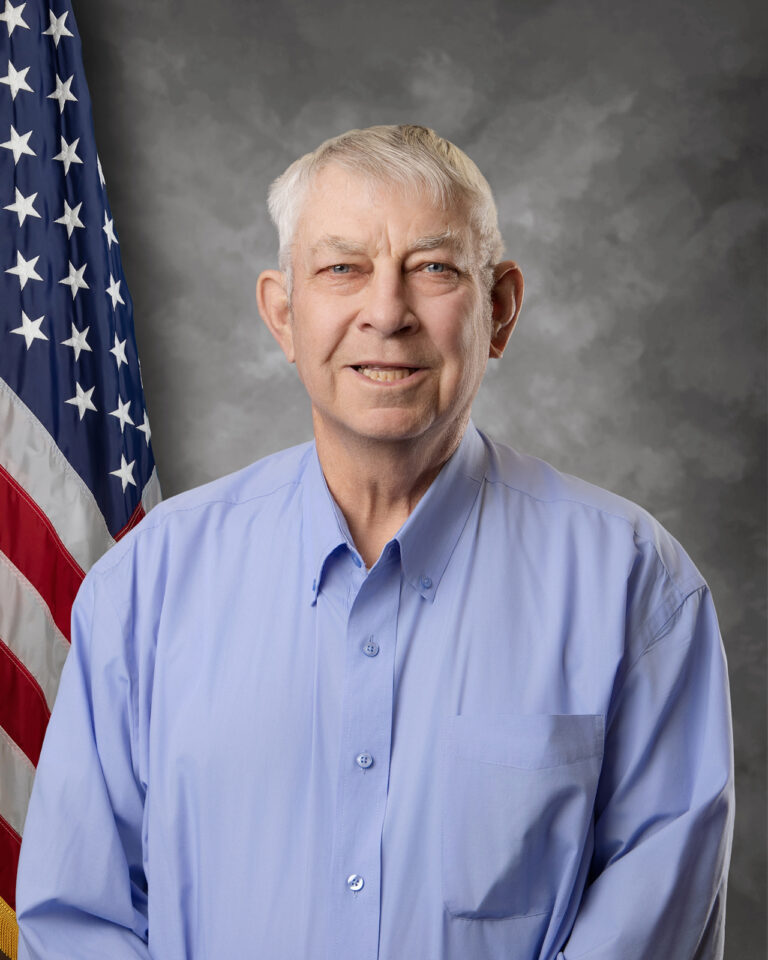 An older man with short gray hair, wearing a light blue button-up shirt, stands in front of an American flag and a gray mottled backdrop, smiling at the camera.