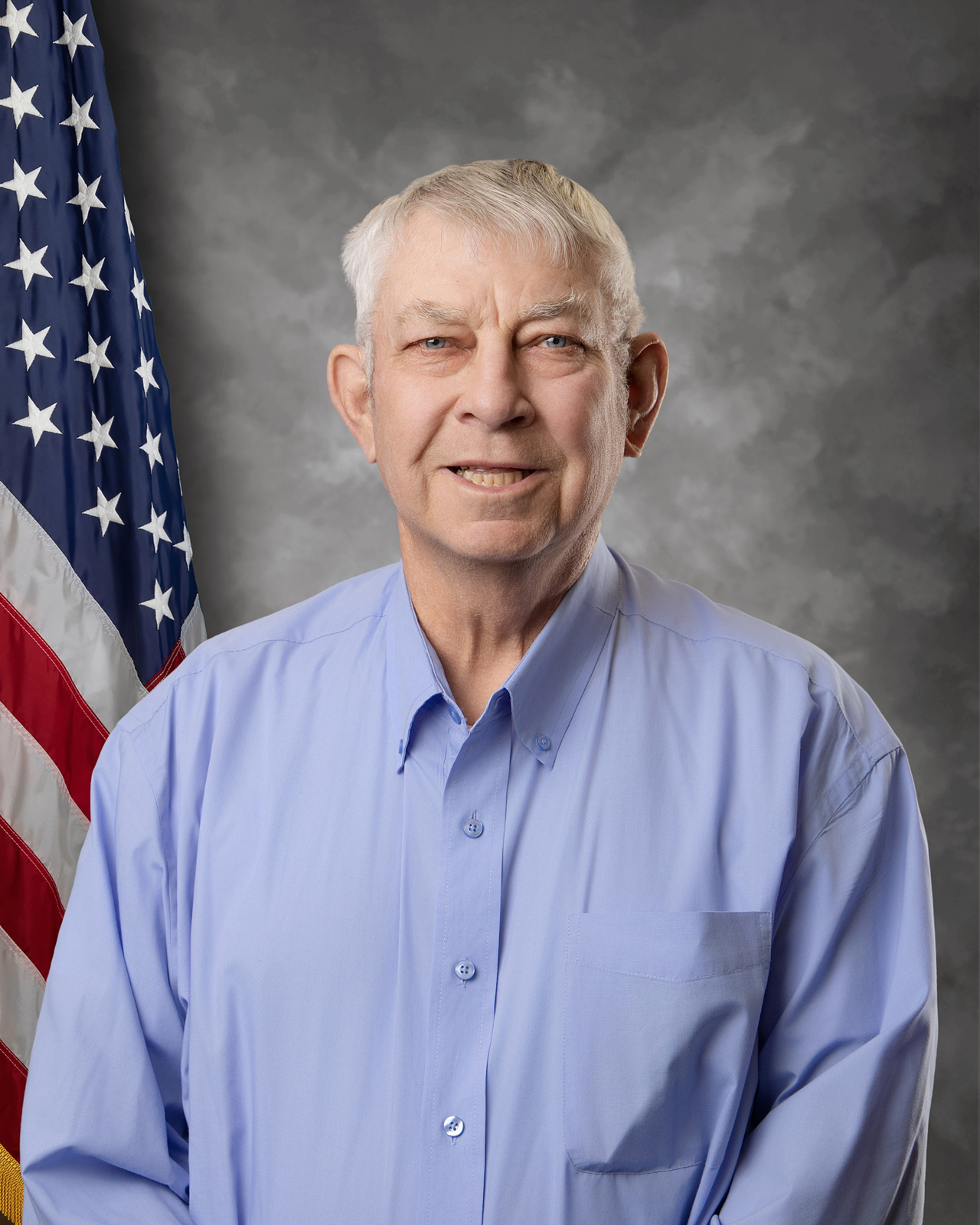An older man with short gray hair, wearing a light blue button-up shirt, stands in front of an American flag and a gray mottled backdrop, smiling at the camera.