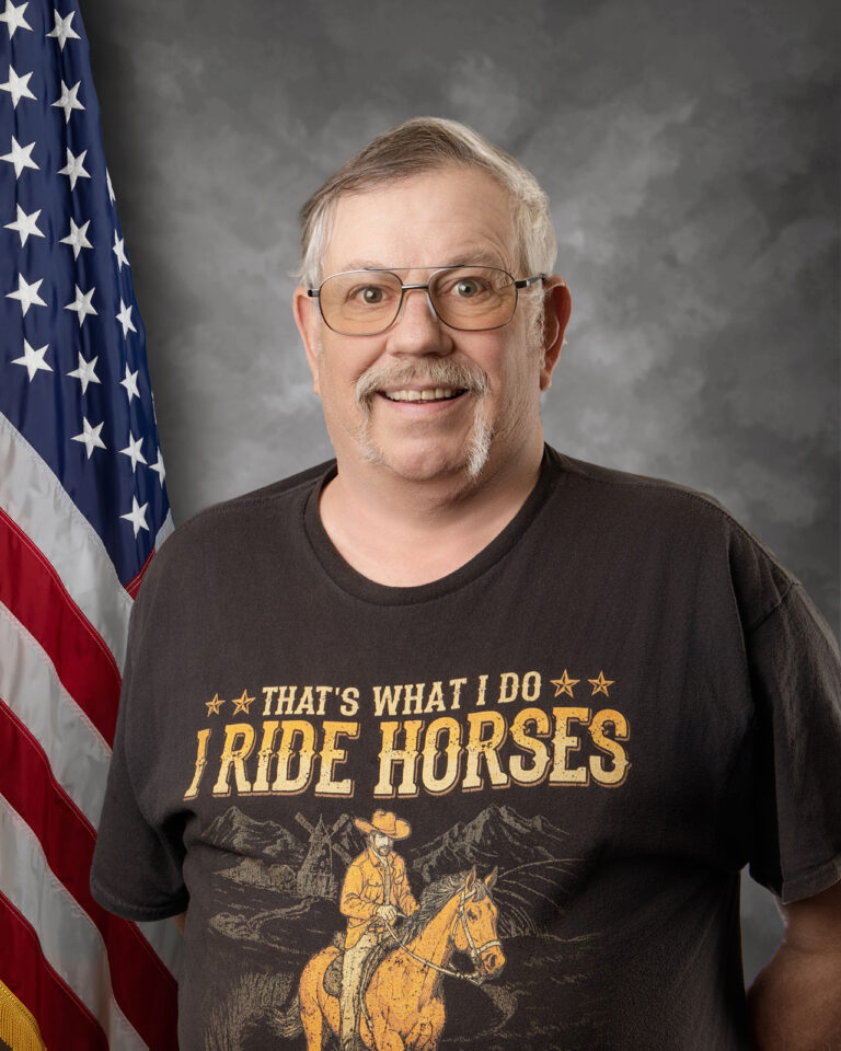 A smiling older man with glasses and a mustache wears a t-shirt that says "That's what I do, I ride horses" with a cowboy graphic, standing in front of an American flag and a gray studio backdrop.