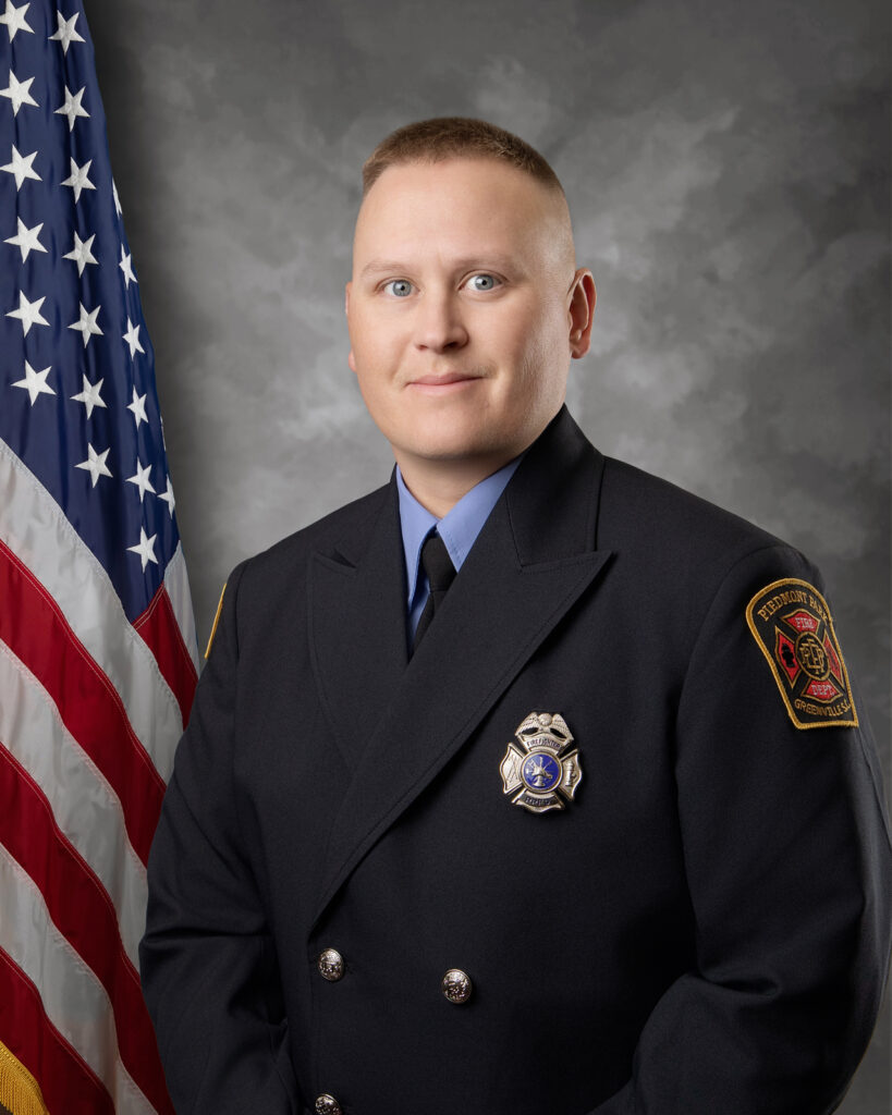 A firefighter in formal dress uniform stands in front of an American flag and a gray backdrop, looking at the camera with a neutral expression. He wears a badge and a patch on his shoulder.