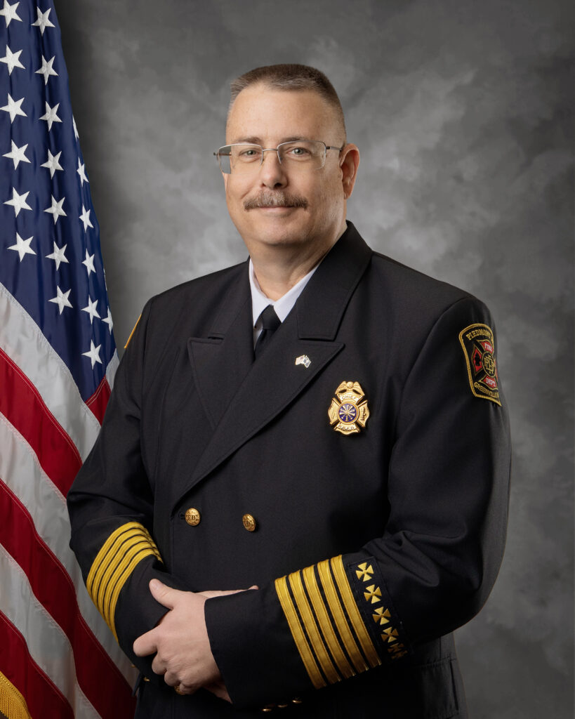 A firefighter in a formal dress uniform stands in front of an American flag, facing the camera with a slight smile. The background is gray and cloudy, creating a formal portrait setting.