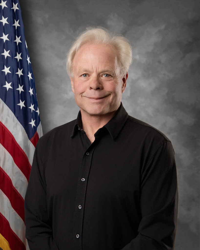 A man with white hair wearing a black button-up shirt stands in front of a cloudy gray background with a United States flag visible on the left side.