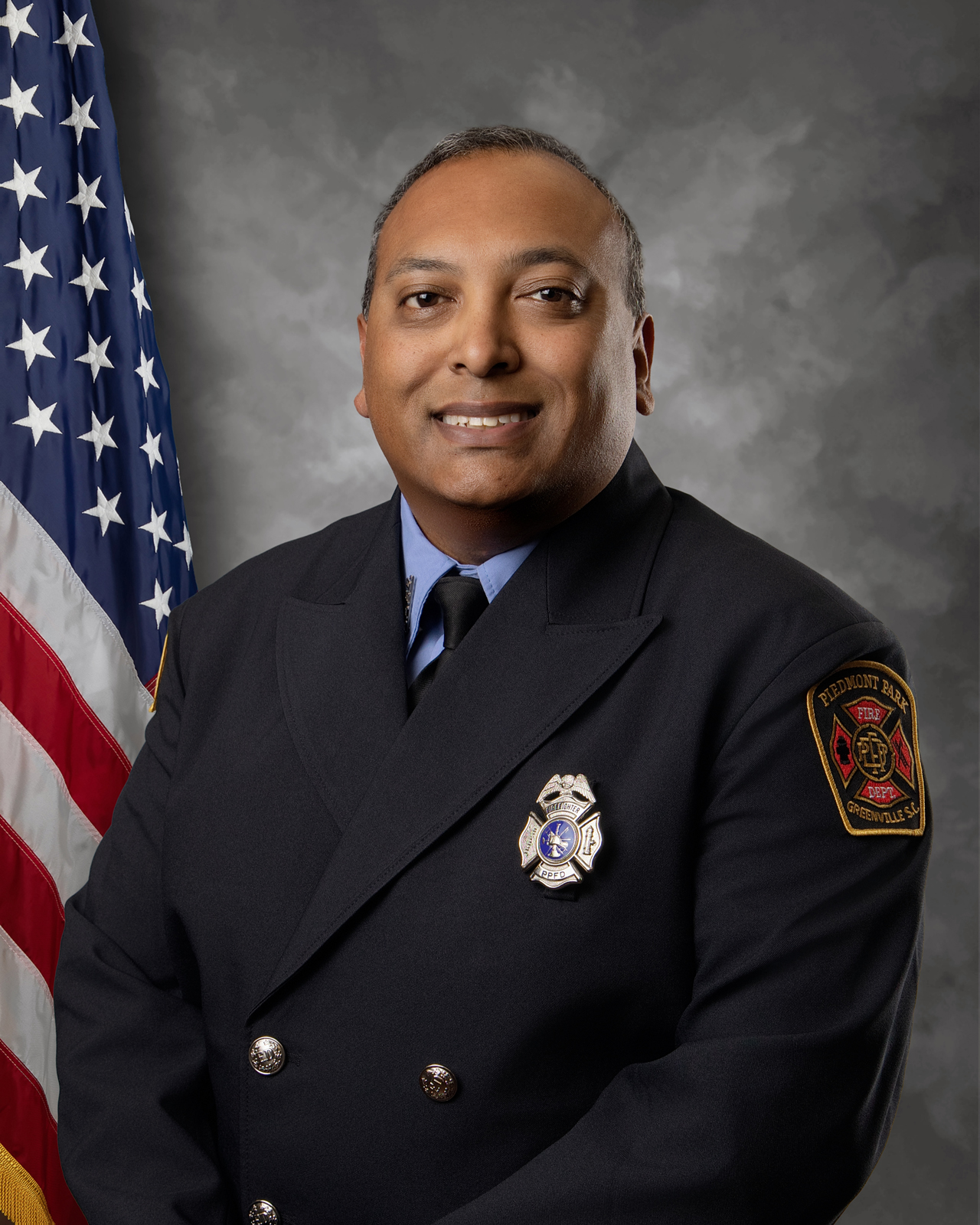 A firefighter in formal dress uniform poses in front of a U.S. flag. He is smiling, wearing a dark jacket with badges and an insignia patch on his upper arm, against a gray studio background.