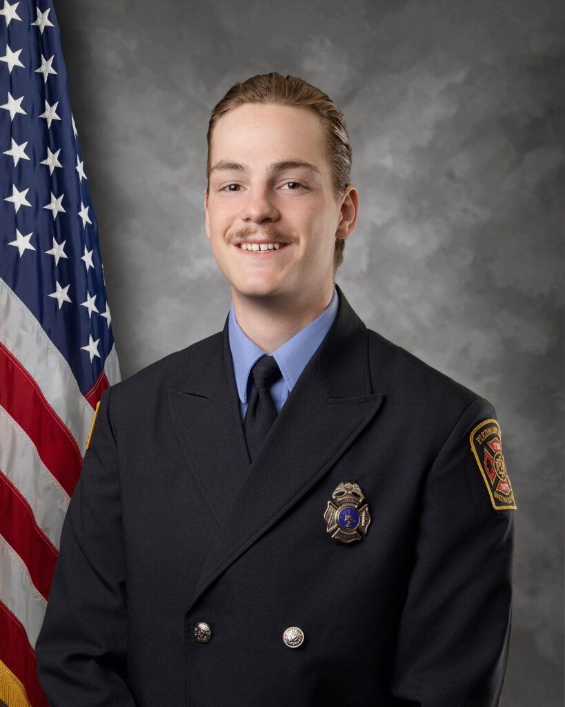 A young firefighter in uniform poses for a formal portrait in front of an American flag and a grey background, smiling slightly at the camera.