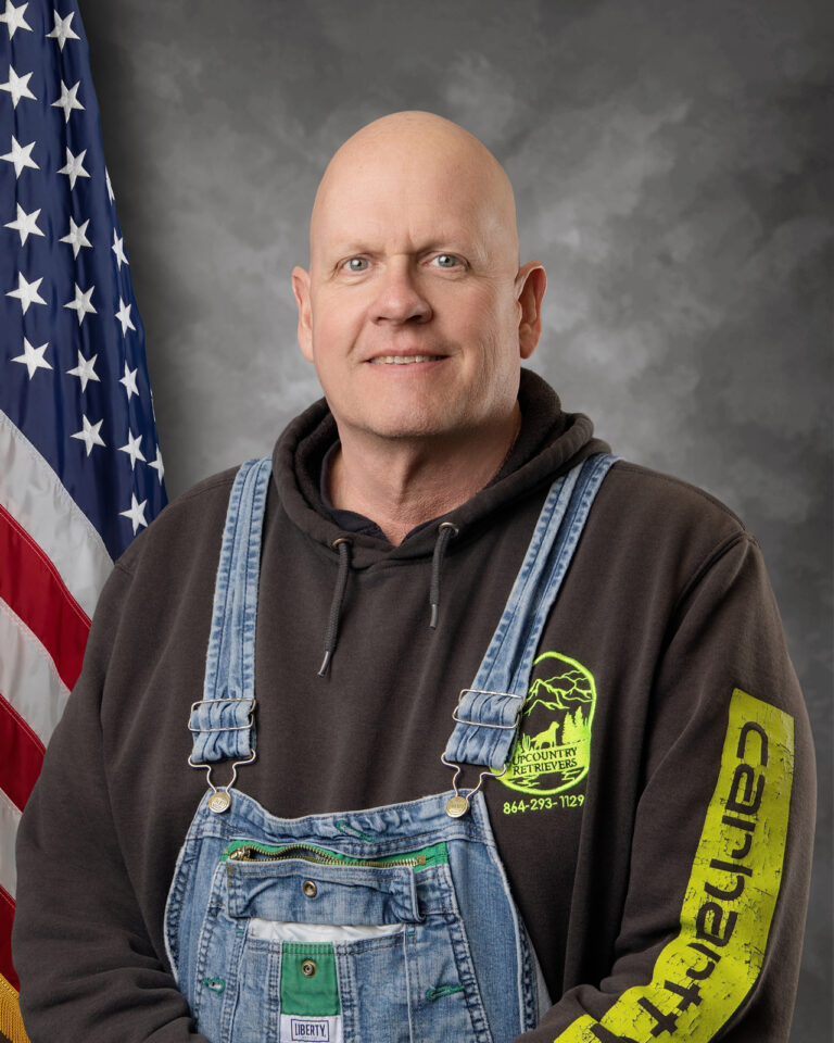 A bald man wearing a dark hoodie with a neon green logo and denim overalls stands in front of an American flag and a gray mottled background, smiling slightly at the camera.