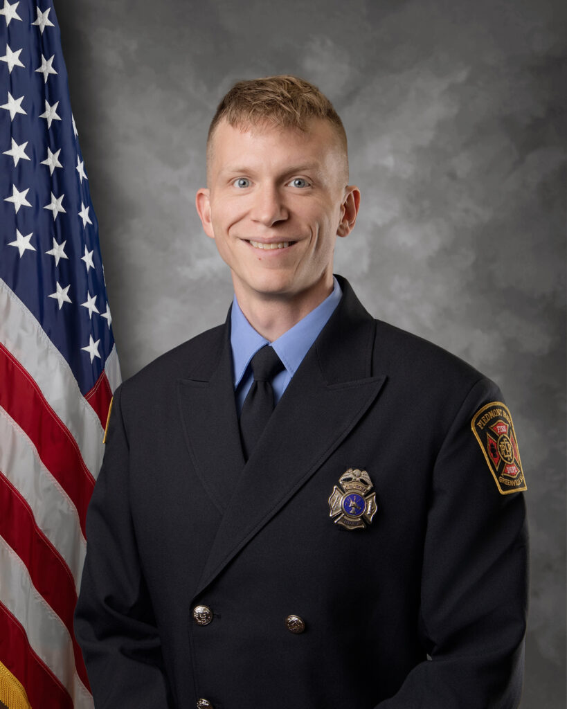 A smiling firefighter in a formal dark uniform poses in front of a U.S. flag and a neutral gray background. The uniform features a badge and a patch on the sleeve.