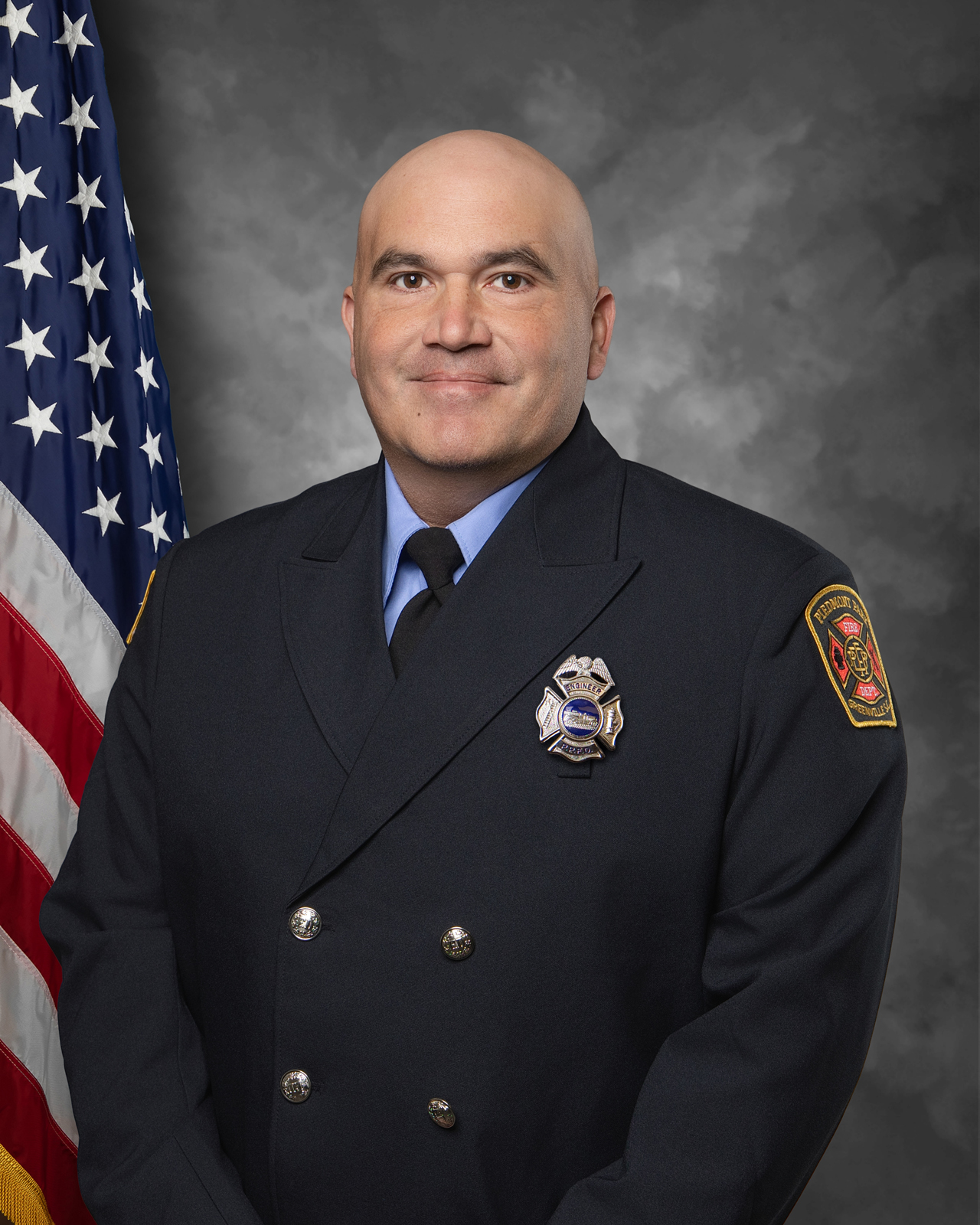 A firefighter in uniform stands in front of an American flag, smiling, with a fire department badge on his jacket and a fire department patch on his sleeve. The background is a neutral gray.