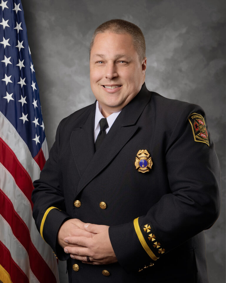 A man in a dark formal dress uniform with gold accents stands in front of an American flag, smiling, with his hands clasped. A fire department badge and patch are visible on his jacket.